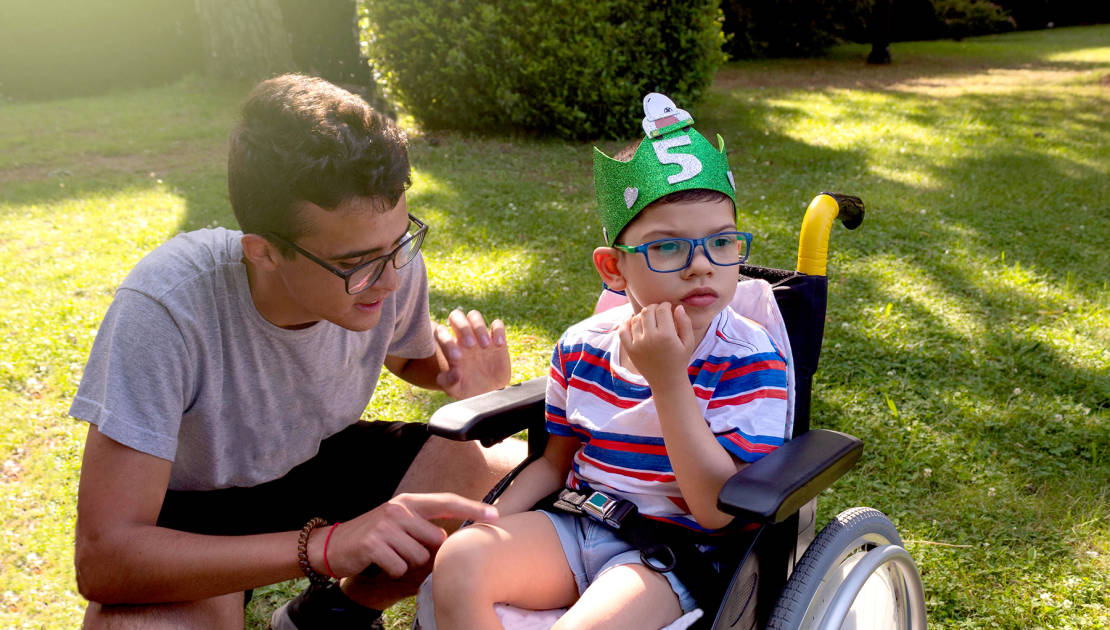 child in wheelchair with Pitthopkins syndrome celebrating his birthday in a garden with a handmade eva rubber wreath and a teenager looking after him