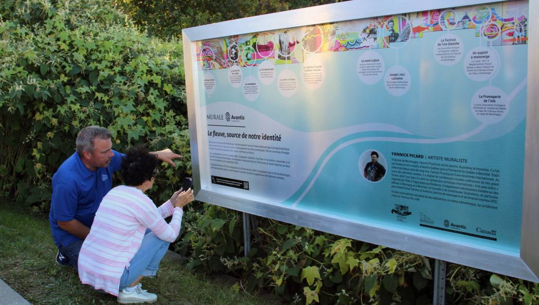 Des visiteurs devant l’affiche explicative de l’œuvre photographiant le code QR pour avoir plus d’informations sur les symboles historiques de la murale. (Photo de courtoisie)