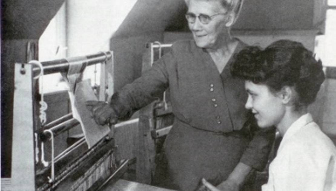 Émélie Chamard donnant des instructions à une tisserande lorsque les métiers à tisser étaient à l’étage de la maison familiale, à Saint-Jean-Port-Joli, vers 1967. (Crédit: Maurice Marquis)